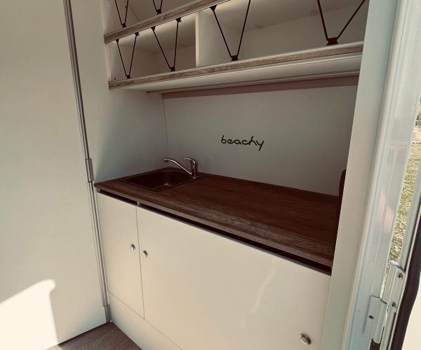 Kompakte und helle Küchenzeile in einem Wohnmobil mit Holzarbeitsplatte, Edelstahlspüle und weißen Unterschränken. Über der Spüle steht "beachy" geschrieben.