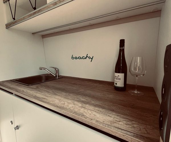 Kompakte und helle Küchenzeile in einem Wohnmobil mit Holzarbeitsplatte, Edelstahlspüle und weißen Unterschränken. Über der Spüle steht "beachy" geschrieben.