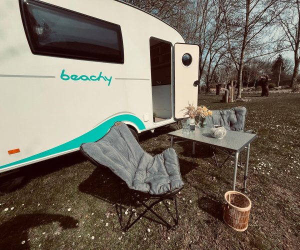Weißer Beachy-Wohnwagen mit türkisfarbenen Akzenten und dem Schriftzug "beachy", davor ein aufgeklappter grauer Campingstuhl und ein Campingtisch mit Blumen, Gläsern und einer Dekoschale.